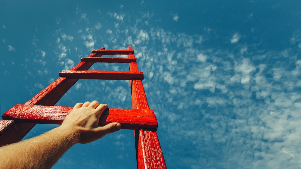 A male hand holds onto the crossbar of a red wooden staircase leading to the blue sky