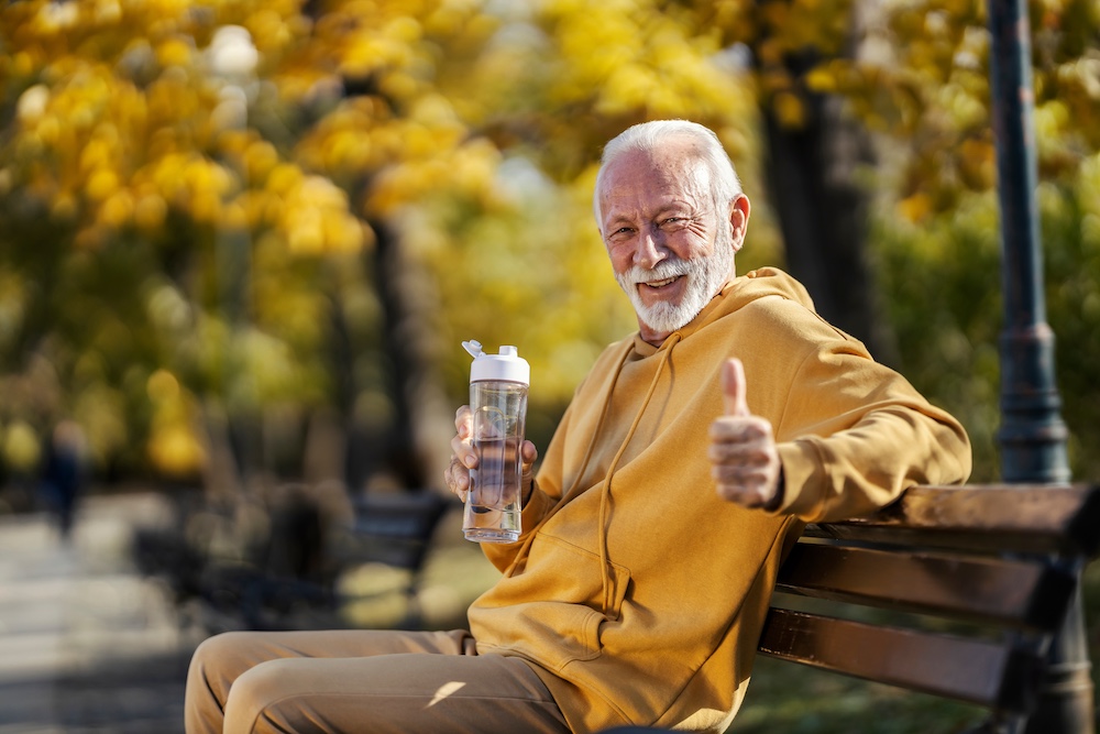 Portrait of senior sportsman sitting on a bench in park with bottle of water in hands and giving thumbs up at camera.